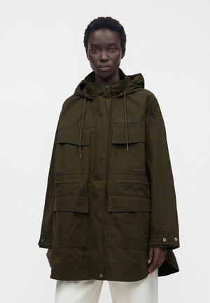 Personne portant une veste à capuche oversize vert olive avec quatre poches à rabat sur le devant et un pantalon blanc, debout devant un fond uni.