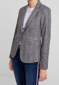 Grauer Blazer mit vertikalen rosa und braunen Streifen, strukturierten Revers, zwei vorderen Taschen und einem Knopfverschluss, getragen über einem helllila Top.