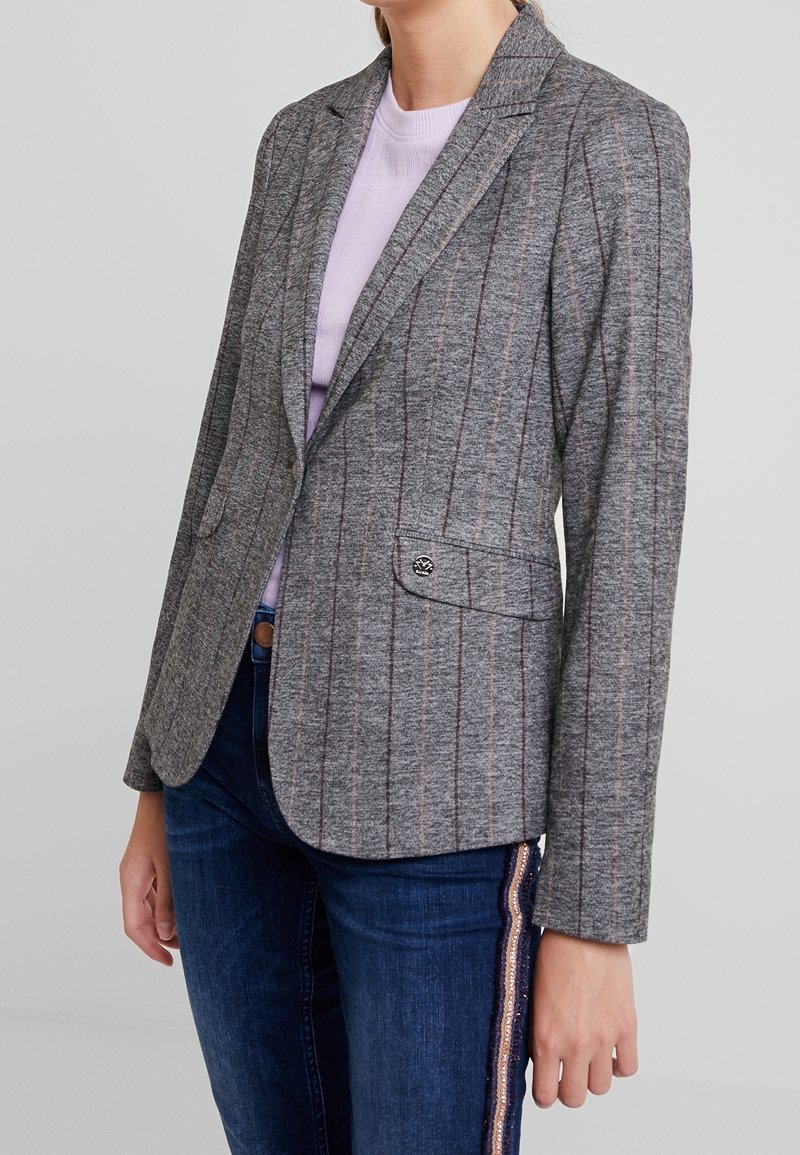 Grauer Blazer mit vertikalen rosa und braunen Streifen, strukturierten Revers, zwei vorderen Taschen und einem Knopfverschluss, getragen über einem helllila Top.