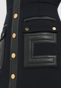 Vestido de tela negra con un bolsillo de acento de cuero con un diseño geométrico y botones de herrajes dorados en la parte delantera para abrochar.
