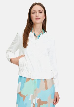 White zip-up jacket with eyelet texture, elastic waistband, and two side pockets, worn over a multicolored patterned dress.
