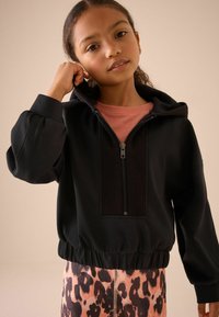Hoodie noir zippé avec un panneau texturé, ourlet resserré et manches bouffantes, associé à des leggings à imprimé léopard rose.