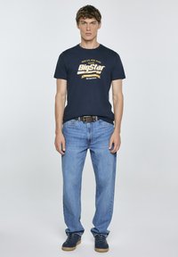 Jūrinės medvilnės marškiniai su geltonu grafikiniu tekstu, derinami su šviesiai mėlynomis džinsinėmis kelniomis. Modelis dėvi tamsius sportinius batelius ir juodą diržą.