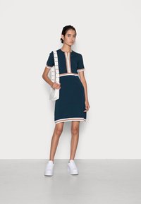 Vestido azul marino hasta la rodilla con mangas cortas, ribetes en blanco y rosas, y cremallera frontal. Combinado con zapatillas de plataforma blancas y un bolso blanco.