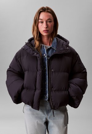 Kvinde iført en sort dunjakke over en blå denimjakke og lysegrå jeans, der står mod en ensfarvet grå baggrund.