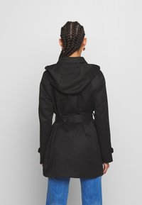 Černý trenčkot s kapucí, páskem v pase a knoflíkovými detaily. Hladká textura, délka ke kolenům, zadní klín pro pohyb.