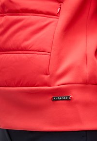 Λεπτομέρεια κόκκινου μπουφάν Calvin Klein που δείχνει μια πλαϊνή τσέπη με φερμουάρ και ένα μικρό μεταλλικό λογότυπο της μάρκας κοντά στο τελείωμα.