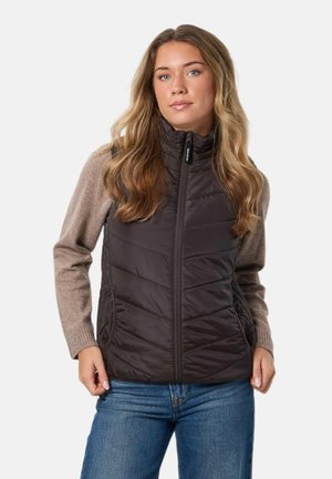 Femme portant un gilet noir matelassé à fermeture éclair sur un pull taupe et un jean bleu, sur un fond clair uni.