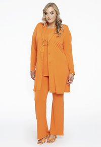 Oranges Ensemble bestehend aus einem langen Jacket, Top und Hose aus glattem Stoff; umfasst gelbe Absatzsandalen und eine runde Halskette als Akzent.