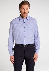 Camicia a righe blu e bianche con maniche lunghe, taschino sul petto e bottoni neri, abbinata a pantaloni scuri.