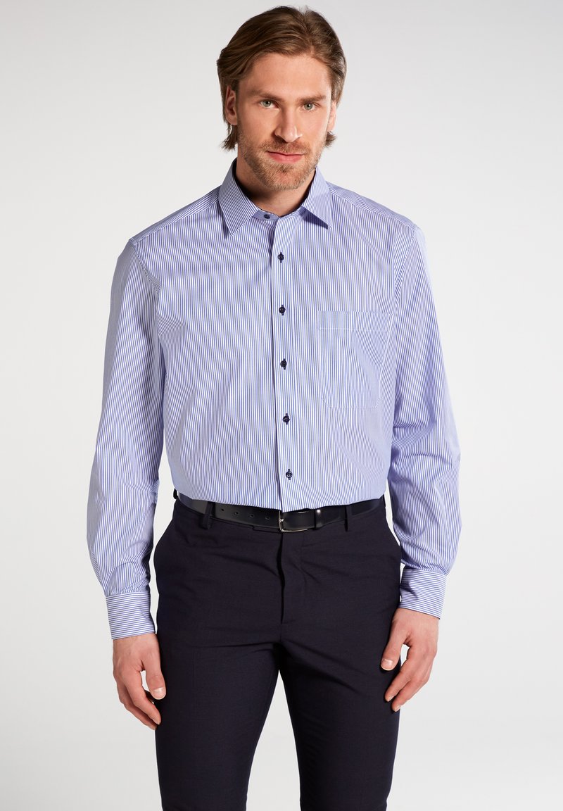 Camicia a righe blu e bianche con maniche lunghe, taschino sul petto e bottoni neri, abbinata a pantaloni scuri.
