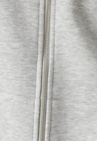 Primer plano de una tela gris claro con un zipper metálico centrado y detalles de costura visibles en la costura.