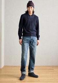 Pullover a maglia blu navy con colletto zip, jeans blu e scarpe scure. Include un berretto a righe blu navy e rosso. Outfit semplice e informale.