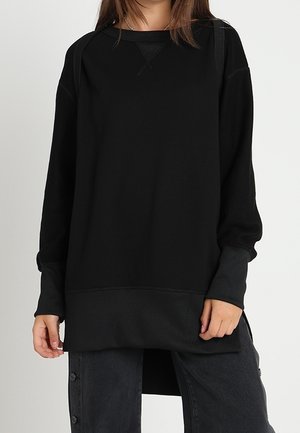 Vrouw draagt een oversized zwart sweatshirt met lange mouwen en donkere broek met knoopdetails aan de zijnaden, staand tegen een effen achtergrond.