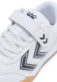 Chaussure de sport blanche avec une tige en mesh, des lacets plats et une sangle à crochets. Accents en zigzag noir sur le côté et semelle en caoutchouc gomme.