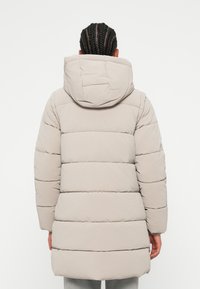 Personne avec des cheveux tressés portant un manteau léger en duvet beige clair avec une capuche, vue de dos sur un fond uni.