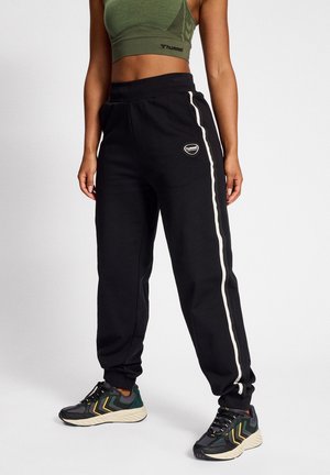 Femme portant un jogging noir avec des bandes blanches sur les côtés, des baskets noires et vertes, et un crop top vert olive, debout sur un fond blanc.