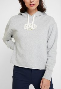 Sudadera corta gris hecha de algodón suave, con un logo bordado blanco de "GAP" y detalles amarillos, además de capucha con cordones.