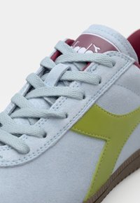 Light blue suede sneaker with green accent, textured laces, red lining, and a logo tag. Features a rubber sole. Close-up view.