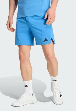 Man draagt blauwe sportbroek, witte Adidas-sokken en witte Adidas-sneakers, staand tegen een effen achtergrond.