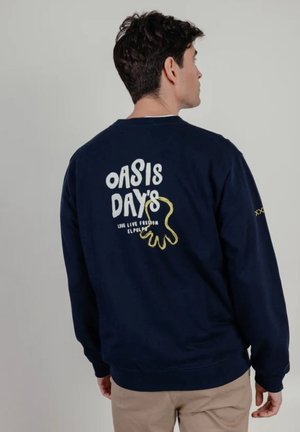 Hombre de espaldas vestido con sudadera azul marino con texto blanco "OASIS DAYS" y diseño abstracto amarillo en la parte trasera, pantalones beige.