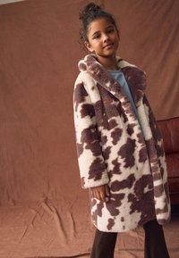 Płaszcz z futra syntetycznego w brązowy i kremowy wzór przypominający skórę krowy, z kapturem, miękką teksturą i długimi rękawami. Noszony na niebieskiej bluzce.