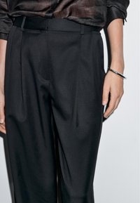 Czarne dopasowane spodnie charakteryzują się gładką teksturą, przednimi fałdami i bocznymi kieszeniami. Na nadgarstku znajduje się srebrna bransoletka.