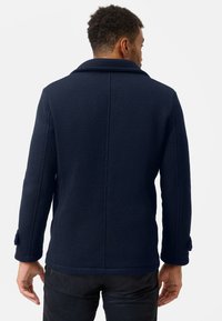 Mörkblå texturerad kappa med bred krage, sydda detaljer och knäppningar på ärmarna, som visar en ren bakvy.