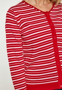 Punainen ja valkoinen raidallinen neule cardigan, jossa on V-aiheinen kaula-aukko ja nappisuljenta, sekä ribbiohennelma ja pitkät hihat.