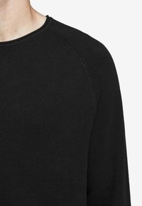 Pull noir en maille avec un tissu texturé, un col rond et des manches raglan. Le matériau semble doux et léger.