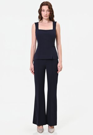 Femme debout portant un haut noir sans manches à encolure carrée et un pantalon large assorti avec des talons hauts beiges.
