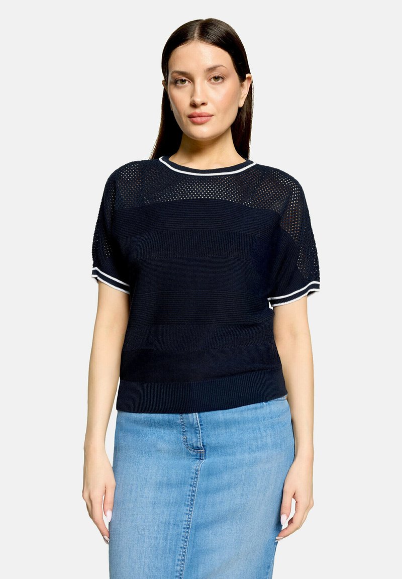 Femme portant un haut en maille noir à manches courtes avec bordure blanche et une jupe en jean bleu clair, face à un fond clair.