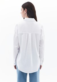 Weißes Langarmhemd mit einer Rückenpasse, knöpfbaren Manschetten und einem abgerundeten Saum. Leicht durchscheinender Stoff; getragen mit blauen Denim-Hosen.