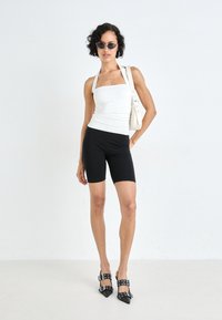 Bela oprijeta halter majica, črne kolesarske kratke hlače do kolen, črni čeveljčki s špičastim podplatom in zaponkami ter velike sončna očala. Nevtralno ozadje.