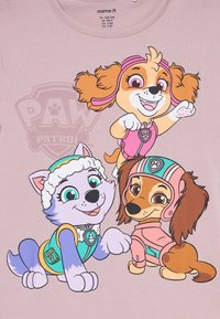 Trois chiens animés de Paw Patrol portant des tenues colorées et des casques sur un fond rose clair.