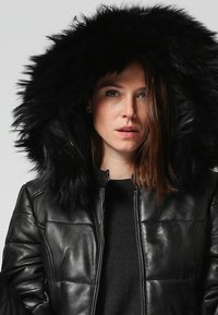 Giacca in pelle nera con un design trapuntato e un ampio cappuccio foderato in pelliccia sintetica. Presenta una zip frontale e polsini a costine.
