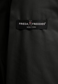 Czarna tkanina o gładkiej fakturze ma prostokątną metkę z napisem "FRIEDA FREDDIES NEW YORK", ozdobioną małym logo psa.