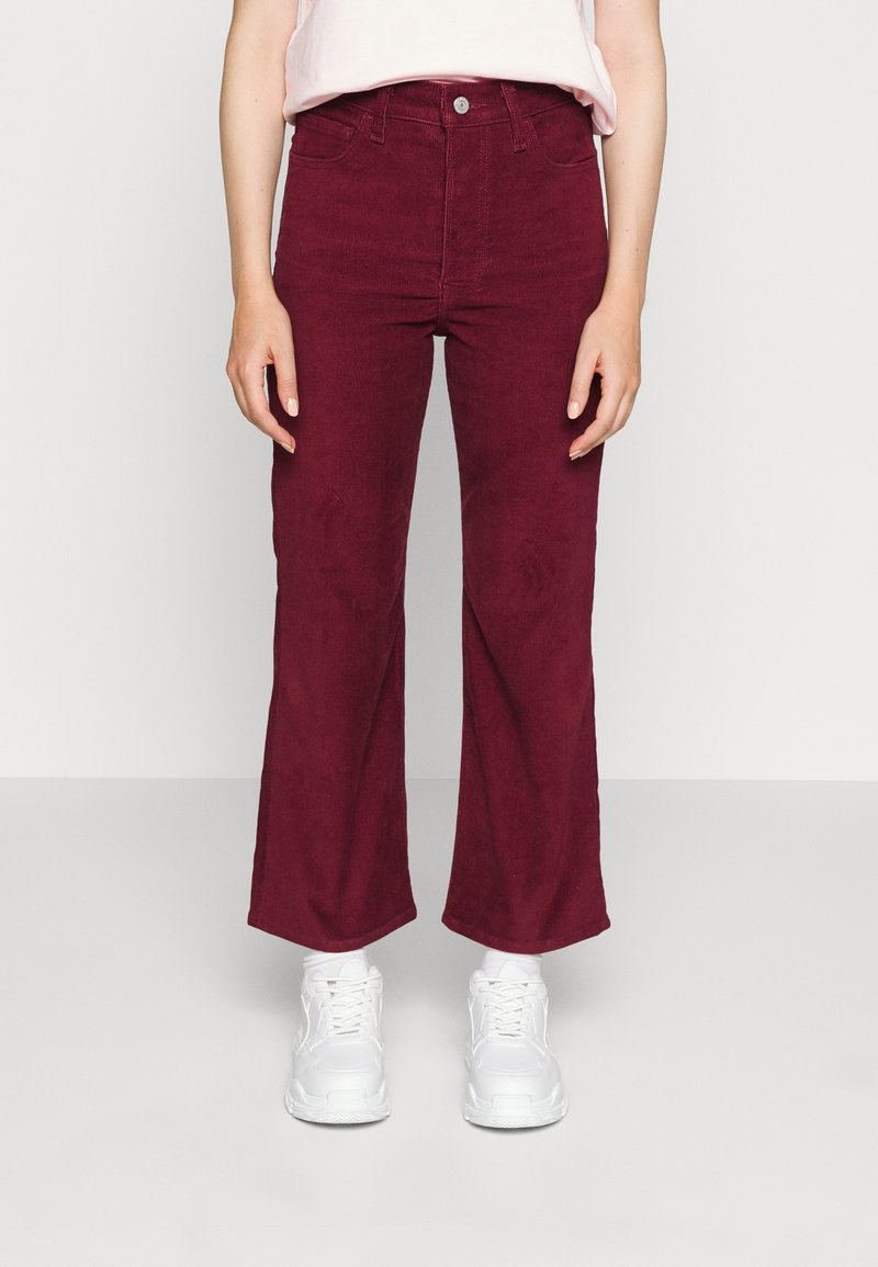 Burgundyröda vida byxor gjorda av mjuk bomull med framfickor, försedda med knapp- och dragkedjestängning, i kombination med vita sneakers.