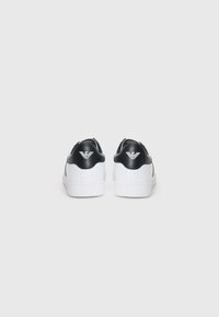 Adidași albi cu accente din piele neagră și un logo pe călcâi. Textură netedă, vârf rotund, guler căptușit și talpă din cauciuc albă.