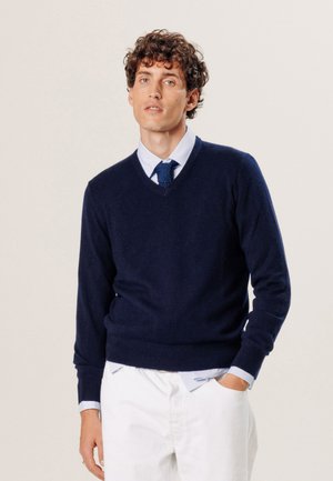 Marineblauwe wollen v-hals trui met geribbelde manchetten en zoom. Draag het over een lichtblauw overhemd met kraag, gestyled met witte broek. Eenvoudig ontwerp.