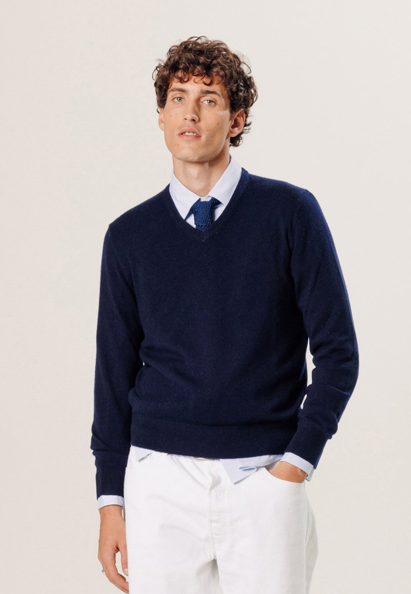 Pull en laine bleu marine avec un col en V, doté de bords côtes aux poignets et à l'ourlet. Porté par-dessus une chemise à col bleu clair, associé à un pantalon blanc. Design simple.