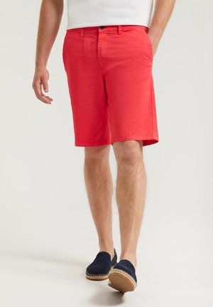 Shorts en coton rouge avec une coupe droite, des poches latérales et une fermeture à bouton, associés à des espadrilles bleues marine. Texture lisse et style ajusté.