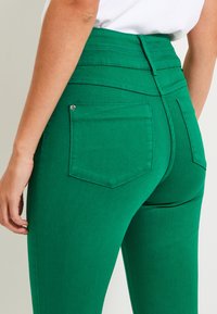 Primo piano di una persona che indossa pantaloni verdi a vita alta con tasche posteriori e una maglietta bianca, mostrando la parte bassa della schiena e la mano appoggiata di lato.