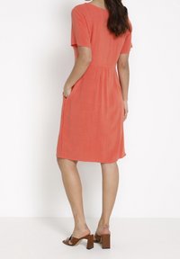 Korallfärgad klänning i knälängd, tillverkad av lätt material, med korta ärmar, fickor och tight midja, kombinerad med bruna sandaler med blockklack.