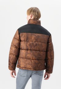 Chaqueta de plumas marrón con estampado floral, hombros acolchados negros y cuello alto. Presenta una forma cuadrada y secciones cosidas para aislamiento.