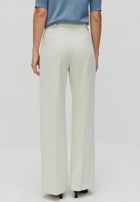 Vida byxa i krämfärgat tyg; mjuk textur, två bakfickor, skräddarsydd design, kombinerat med en blå topp, stilrena svarta klackar.