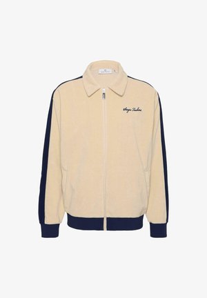 Kremfarget jakke med glidelås og marineblå sidepaneler, laget av mykt stoff. Har krage, ribbede mansjetter og brodert logo på brystet.