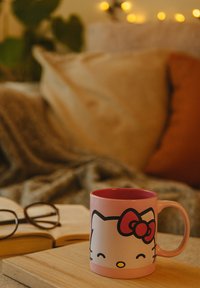 Pink Hello Kitty mug on wooden table with open book and glasses, cozy couch and string lights in the background.
