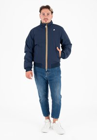 Giacca puffer blu navy con colletto alto, dettaglio con zip gialla e polsini elasticizzati. Indossata sopra jeans blu e sneakers bianche.