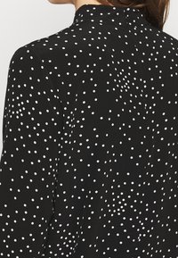 Zwarte blouse met een witte stippenpatroon, voorzien van een hoge kraag en een gladde stoftextuur. Het ontwerp bedekt de rug en schouders.